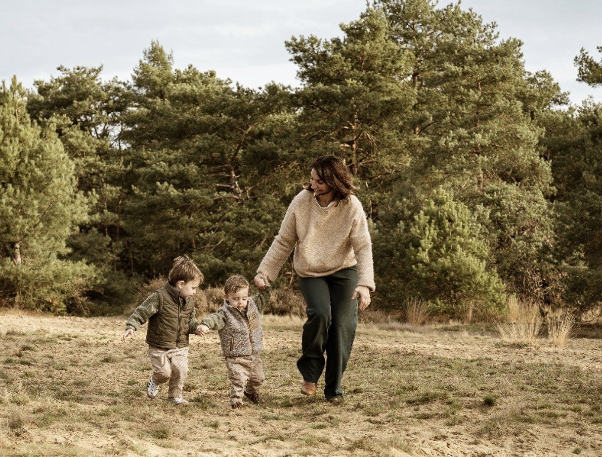 Een vrouw wandelt hand in hand met twee jonge kinderen in een bosrijke omgeving.