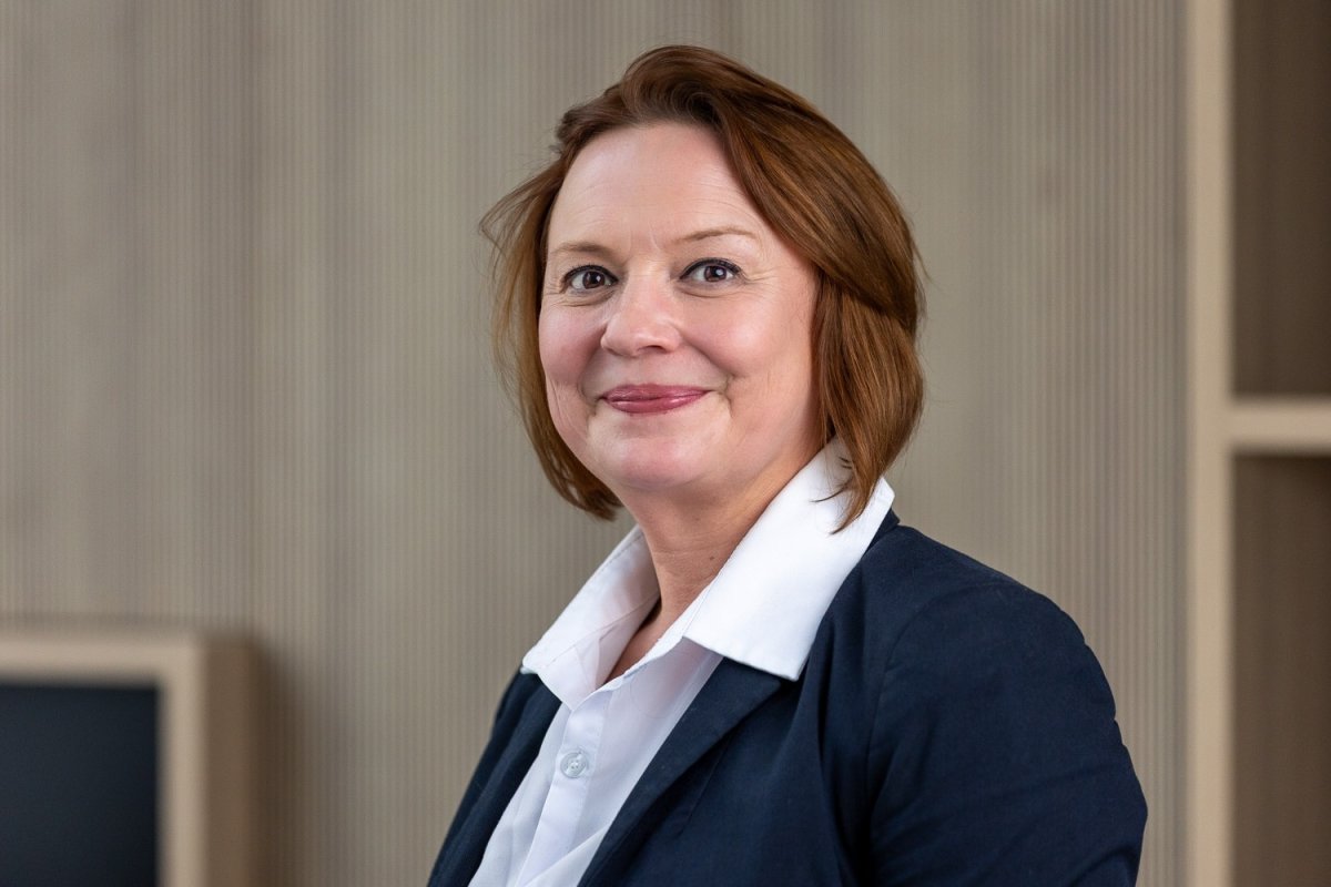 Portret van een vrouw met halflang haar, gekleed in een donkerblauwe blazer en witte blouse, met een glimlach op haar gezicht en een neutrale achtergrond.