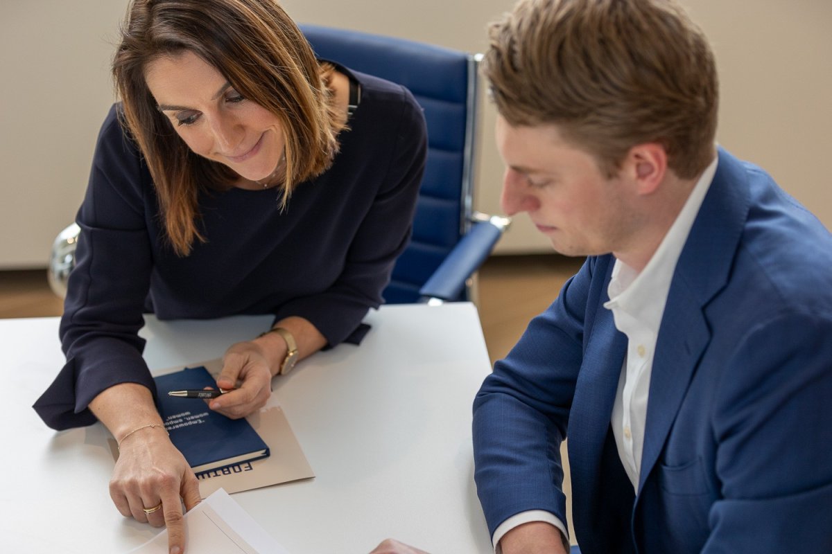 Twee mensen zitten aan een tafel, waarbij een vrouw met bruin haar de aandacht op een document wijst terwijl een man in een blauw jasje naar het papier kijkt.