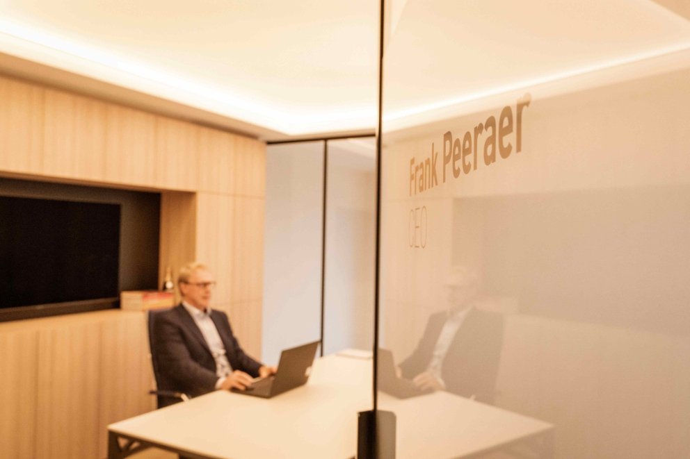 Een man in een blazer zit aan een bureau in een vergaderruimte met een laptop voor zich, met een glazen wand waarop de naam "Frank Peeraer, CEO" staat.