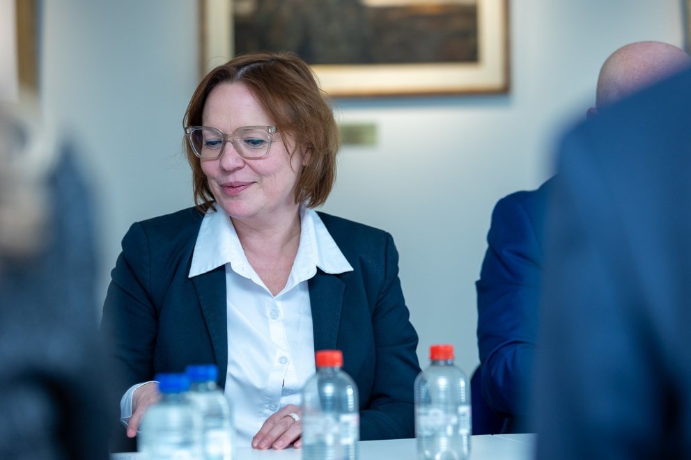 Een vrouw met bruin haar en bril in een zakelijke outfit, zittend aan een vergadertafel met waterflessen, glimlachend en in gesprek.