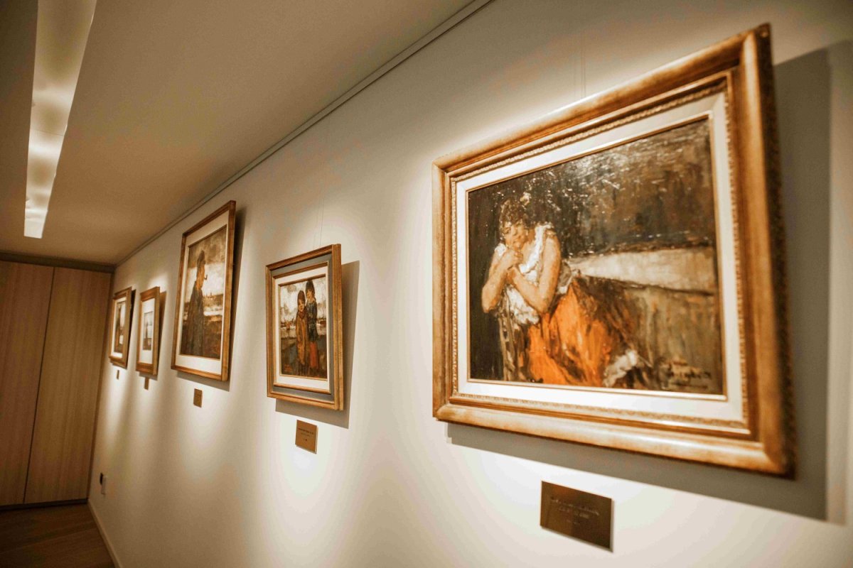 Een galerijwand met vier ingelijste schilderijen, verlicht door een rechte plafondlamp. De schilderijen tonen vrouwen en kinderen in verschillende poses en stijlen.