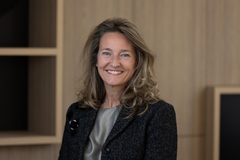 Portret van een vrouw met lang, golvend haar, die vriendelijk glimlacht, gekleed in een donkere jas en een lichtgekleurde blouse, tegen een neutrale houten achtergrond.